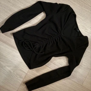 Svart omlottopp med snörning - Snygg svart omlottopp med lång ärm och v-ringning. Toppen har snörning framtill som ger en cool, justerbar look. Perfekt att styla med jeans eller kjol för en trendig outfit.