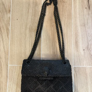 Svart axelväska Kurt Geiger  - Snygg svart axelväska från Kurt Geiger. Väskan har kedjerem, dekorativa svarta paljetter i rutmönster och ikoniskt örnhuvud på locket. Perfekt storlek för det viktigaste och ger en cool vibe till din outfit.