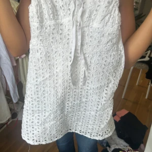 Vit klänning  - Söt vit klänning  i bomull med broderade hålmönster och knyt framtill. Ärmlös modell med lätt A-linjeformad passning, perfekt för varma dagar. Ger en fräsch och somrig känsla till din outfit.köpt i Spanien 