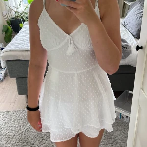 Vit kort sommarklänning med prickar - Supersöt vit kort klänning med tunna axelband och v-ringning. Klänningen har ett diskret prickigt mönster oxj ett par inbyggda shorts som gör klänningen mer bekväm.