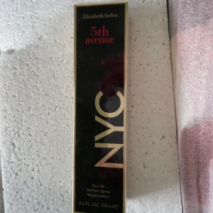Elizabeth Arden 5th Avenue NYC Parfym - Snygg parfym från Elizabeth Arden, 5th Avenue NYC. Flaskan rymmer 125 ml Eau de Parfum Spray. Förpackningen är svart med guldiga och röda detaljer, vilket ger en lyxig och modern känsla.