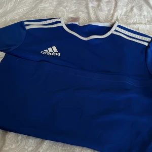 Blå Adidas fotbollströja AEROREADY - Snygg blå fotbollströja från Adidas med klassiska vita ränder på axlarna och vit kant vid halsen. Tillverkad i lätt och ventilerande AEROREADY-material som passar perfekt för träning eller match. Adidas-logga i vitt på bröstet. Änvänd många gånger men är i väldigt bra skick!