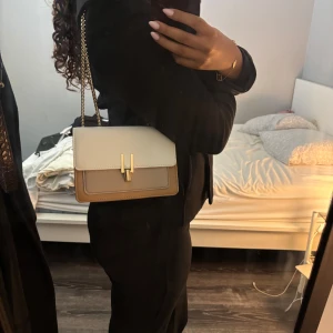 Beige och vit axelväska med guldkedja - Snygg axelväska i beige och vitt med stilren guldkedja och elegant låsdetalj framtill. Väskan har ett innerfack med dragkedja och är perfekt för att bära det viktigaste. Klassisk och clean design som lyfter vilken outfit som helst.