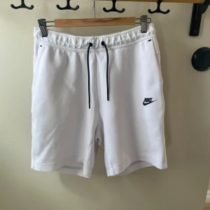 Vita Nike shorts med snörning - Fräscha vita shorts från Nike med svart snörning i midjan och broderad Nike-logga på ena benet. Mjukt material, elastisk midja och praktiska fickor, varav en med dragkedja bak. Perfekta för en sportig look under varma dagar.