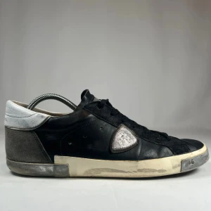 Philippe Model svarta sneakers i skinn - Svarta sneakers från Philippe Model med detaljer i grått och vitt. Ovandel i skinn och mocka, silverfärgad logotyp på sidan och vit/grå sula med vintage-look. Klassisk snörning och låg profil, perfekt för en avslappnad streetstil. Ingen box