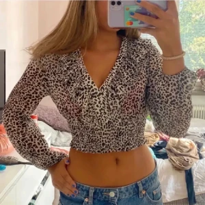 Leopardmönstrad croppad blus - Säljer en croppad blus med leopardmönster i svart och vitt. Toppen har v-ringning, långa ärmar och smockad midja som ger en snygg passform. Perfekt för dig som vill sticka ut med ett trendigt djurmönster.