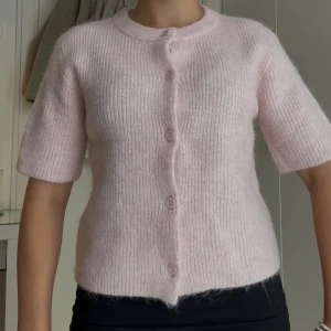 Ljusrosa stickad kofta med knappar 🩷 - Superfin ljusrosa stickad kofta med korta ärmar och rund hals. Koftan har ribbad struktur och stängs med matchande knappar framtill. Perfekt för lager-på-lager och ger en söt, mjuk vibe till din outfit.