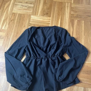 Svart omlottblus med v-ringning - Svart blus med omlott och djup v-ringning, markerad midja och långa ärmar med resår vid handlederna. Snygg spetsdetalj längs ringningen ger en extra touch. Perfekt att styla med jeans eller kjol.