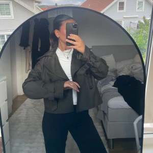 Kort mörkgrön trenchcoat - Säljer en kort trenchcoat från h&m i mörkgrönt med klassiska detaljer som dubbelknäppning, bred krage och bälte vid ärmsluten. Den är i storlek xs men passar xxs-s. Så fin och aldrig använd.