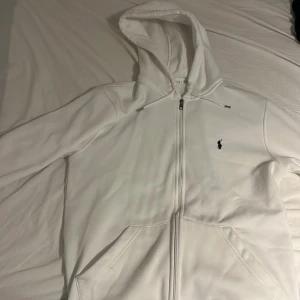 Vit hoodie från Ralph Lauren - Vit hoodie från Ralph Lauren med dragkedja och klassisk blå broderad logga på bröstet. Tröjan har huva med snörning, två fickor framtill och långa ärmar. Perfekt för en clean och stilren look. 