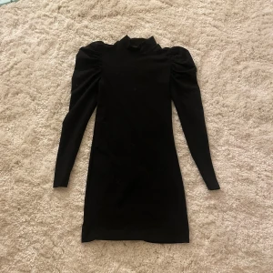 Svart glittrig klänning från Pigalle - Supersnygg svart glittrig klänning från Pigalle med puffade axlar och lång ärm. Hög hals och figurnära passform ger en stilren och trendig look. Perfekt för dig som gillar festligare plagg!🪩