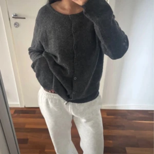 Mörkgrå stickad kofta med knappar - Mysig mörkgrå stickad kofta med rund halsringning och knäppning framtill. Koftan har en enkel och clean design med långa ärmar och en lite loose passform, perfekt att slänga på sig när det är kyligt.