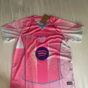 FC Barcelona rosa matchtröja Nike - Snygg FC Barcelona fotbollströja från Nike i rosa och vitt med tunna linjer och klubbmärke på bröstet. Tröjan har korta ärmar, V-ringning och tryckt Spotify-logga på magen. Tillverkad i lätt och ventilerande polyester, perfekt för träning eller match.