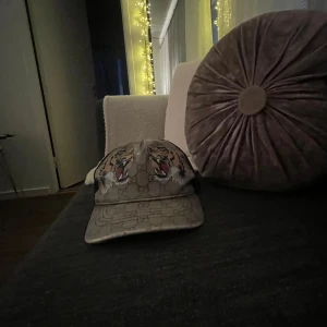  keps med tigertryck - Snygg keps från  med klassiskt -mönster i beige och brunt. Framtill pryds kepsen av två detaljerade tigerhuvuden med röd och svart accent. Baksidan har svart mesh och justerbart spänne. Perfekt för dig som vill sticka ut med en lyxig accessoar.