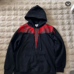  Marcelo Burlon hoddie  - Svart hoodie från Marcelo Burlon County of Milan med fet röd print som liknar vingar över axlar och bröst. Tröjan har huva med snörning, stor magficka och är tillverkad i 100% bomull. Perfekt för dig som vill sticka ut med en unik streetwear-look.