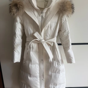 Vit lång dunjacka med pälskrage XS/S - En snygg lång vit dunjacka med fluffig brun äkta pälskrage och bälte i midjan. Jackan har dragkedja framtill, knappar och quiltad design. Perfekt för kalla vinterdagar när du vill hålla stilen och den kommer definitivt att hålla värmen. Endast använd en vinter och ca använd 10ggr. Figursydd i strl XS men passar min strl som är S. Köpt för 4000kr, pris kan diskuteras. Exklusive frakt gäller för varan.