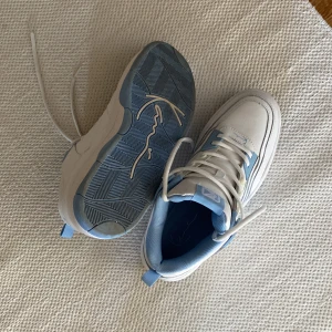 Vit-blå sneakers från Karl Kani - Säljer ett par fräscha sneakers från Karl Kani i vitt och ljusblått. Skorna har en chunky sula, snörning och coolt broderad logga på sidan. Ovandelen är i syntetmaterial med blå detaljer vid snörningen och på insidan. Perfekt för dig som gillar streetstyle.