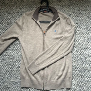 Beige kofta från morris 100% merinoull  - Snygg beige kofta från Morris i 100% merinoull med ribbad mörkbrun krage och hel dragkedja framtill. Liten blå broderad detalj på bröstet. Perfekt för lager-på-lager och chill stil. Passar dig som gillar klassiska och stilrena plagg. Storlek L passar M-L. Nyskick