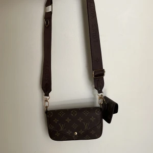 Superfin brun axelväska - Säljer en brun axelväska  med klassiskt monogrammönster och bred justerbar axelrem. Väskan har guldfärgade detaljer och en mindre matchande plånbok som hänger på sidan. Säljer då den inte kommer till användning. 