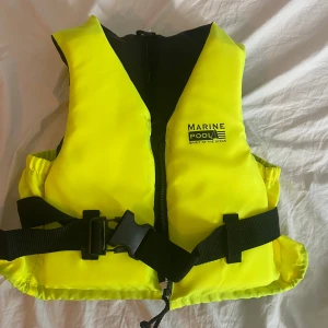 Gul flytväst från Marinepool 30-50kg - Flytväst i neon-gult från Marinepool med svart insida och justerbart midjebälte. Västen har dragkedja framtill och ett stort spänne för extra säkerhet. Perfekt för vattenaktiviteter och syns tydligt tack vare den starka färgen. Passar perfekt för folk som väger 30-50 kg. Andvänt en gång men har bara stått vid land så syns ingenting att den är andvänd. Pris kan diskuteras.