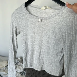 Ljusgrå ribbad topp med spets H&M - Supersöt ljusgrå långärmad topp från H&M med ribbad struktur, små hålmönster och spetskant vid halsen. Söt rosett framtill ger extra detalj. Perfekt för lager-på-lager och chill outfits. Passar som pyjamas och är stretchig! Så passar lite större än 134/140❤️