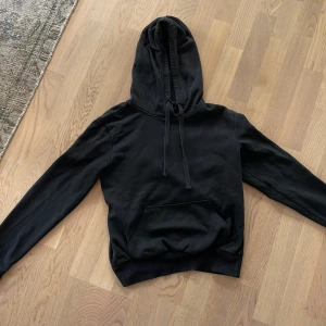Svart hoodie med huva och magficka - En svart hoodie med klassisk huva och snörning, samt en stor magficka framtill. Tröjan har långa ärmar med ribbade muddar och en relaxed passform. Perfekt för chill dagar eller när du vill ha en enkel och stilren look. Den är mjuk och skön. Passar mig som har xs och s i tjock tröjor. 