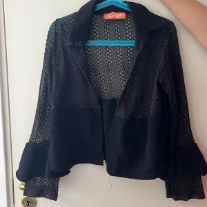 Svart overshirt med spetsdetaljer - Snygg svart overshirt från Inai Kids med genombrutna spetsdetaljer och breda volangärmar. Jackan har krage och är tillverkad i ett lätt, syntetiskt material som ger en luftig känsla. Perfekt för att lyfta vilken outfit som helst.