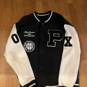 Svart och vit bomberjacka med tryck - Snygg svart och vit bomberjacka med stora bokstäver och broderade detaljer. Jackan har vita ärmar, svart kropp och coola patches med sporttema. Stängs med knappar framtill och har ribbade muddar vid ärmslut och krage.