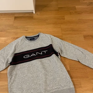 Grå sweatshirt från GANT med logga - Snygg grå sweatshirt från GANT med svart och röd rand över bröstet och stor vit logga. Tröjan har rund hals, långa ärmar och är tillverkad i mjuk bomullsmix. Perfekt för en avslappnad och stilren look.