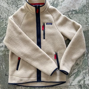 Beige fleecejacka från Patagonia - Säljer en Retro Pile Jacket från Patagonia med marinblå och röda detaljer. Jackan har hög krage, hel dragkedja framtill och tre fickor med dragkedja. Perfekt för kyliga höstdagar!