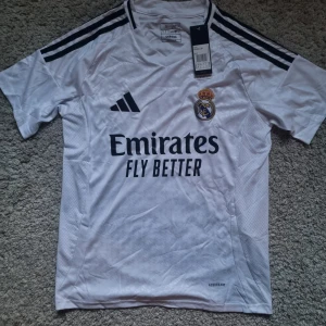 Real Madrid 25 Tröja - Snygg vit Real Madrid fotbollströja från Adidas i slim fit. Klassisk design med svarta ränder på axlarna, klubbmärke på bröstet och 'Emirates Fly Better' tryck framtill. Tillverkad i lätt och ventilerande Aeroready-material, perfekt för match eller träning.