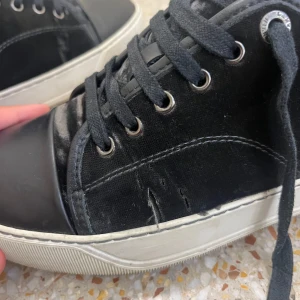 Svarta sneakers från Lanvin -  Pris kan diskuteras. Svarta sneakers från Lanvin med vit platt sula och rund tå. Ovandelen är i mocka med detaljer i skinn på tån och snörning med svarta skosnören. Metallöglor och diskret Lanvin-logga på sulan. Snygg och stilren design som passar till det mesta. . Med lite slitningar vid sidan och knytsnören. Storlek 39-40 Kommer med en dustbag.