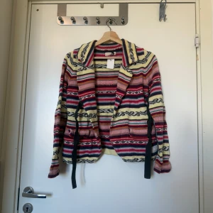 Randig cardigan kofta från Odd Molly - Färgglad kofta från Odd Molly med randigt mönster i rött, rosa, gult, blått och svart. Stickad i mjukt material med krage, knytband i midjan och broderad logga på ryggen. Perfekt för dig som vill sticka ut med en unik och cozy look. Storlek 2 som enligt deras storleksguide motsvarar en storlek M. Fint skick, men kan vara något mer använd. Kan ha några små tecken på användning. 