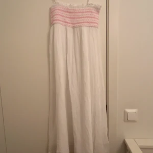 Vit långklänning med rosa broderi - Söt långklänning i vitt med smock upptill och tunna axelband. Överdelen har broderade rosa detaljer och volangkant. Man kan ta bort banden, den har en fläck som ma ser på bilder. 
