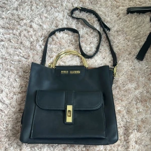 Svart axelväska från Steve Madden - Snygg svart axelväska från Steve Madden med guldfärgade detaljer och handtag. Väskan har en framficka med lås, justerbar axelrem och rymligt foder med innerfack och dragkedja. Perfekt för dig som vill ha både stil och funktion.