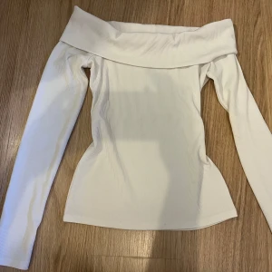 Vit offshoulder ribbad topp - Säljer en stilren vit offshoulder topp med långa ärmar och ribbad struktur. Toppen har en bred, vikt kant upptill som ger en snygg och modern look. Perfekt för dig som gillar minimalistisk och trendig stil. Den är från hm.