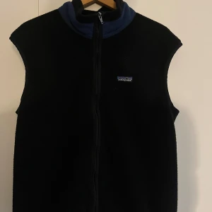 Patagonia väst - Svart fleeceväst från Patagonia med blå krage och hel dragkedja framtill. Klassisk och enkel design med liten logotyp på bröstet. Perfekt lager-på-lager-plagg för kyliga dagar. Storlek L men passar även M