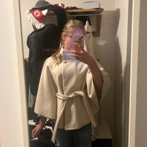 Beige kappa med bälte från Missay - Säljer en beige kappa från Missay med snyggt bälte i midjan och hög krage. Jackan har en elegant, loose passform och vida ärmar som ger en cool siluett. Perfekt för dig som gillar stilrena och trendiga plagg. Knappar framtill för enkel stängning.