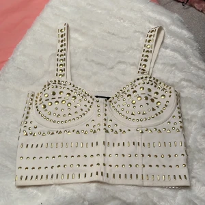 Vit korsettopp med guldnitar - Supersnygg vit korsettopp med breda axelband och massor av guldiga nitar i olika former över hela framsidan och banden. Toppen har markerade kupor och en croppad passform som ger en edgy vibe. Perfekt för dig som vill sticka ut och älskar detaljer!