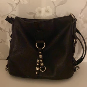 Svart axelväska med nitar och mönstrat foder - Cool svart axelväska i skinn med silverfärgade nitar och ringdetaljer framtill. Väskan har en bred axelrem och dragkedja baktill. Insidan är fodrad med ett snyggt svartvitt blommigt mönster. Perfekt storlek för allt du behöver ha med dig. har 2 små fläckar på insidan men det är rent bara att de inte går bort.