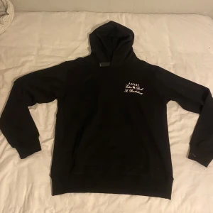 Svart Amiri hoodie med tryck - Svart hoodie från Amiri med vit text och motiv både fram och bak. På bröstet står det 'AMIRI Eden Rock St. Barthelemy' och på ryggen är det ett stort tryck med samma text och ett flygplansmotiv. Klassisk känguruficka och huva. Perfekt för dig som gillar streetwear.