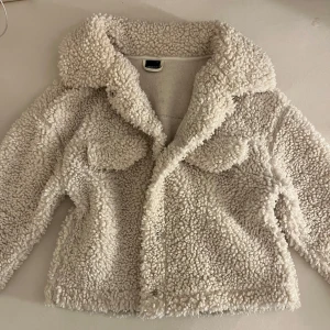 Vit teddyjacka från Gina Tricot - Supermysig vit teddyjacka från Gina Tricot i storlek S. Jackan har stora fickor framtill, bred krage och stängs med tryckknappar. Perfekt för att hålla sig varm och snygg under kyliga dagar. Jackan har en fluffig och trendig look som passar till allt.