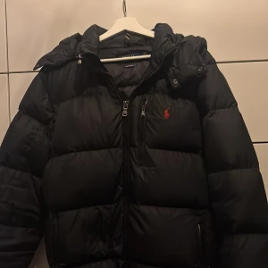 Svart pufferjacka från Polo Ralph Lauren - Svart pufferjacka från Polo Ralph Lauren med ikonisk röd logga på bröstet. Jackan har huva, flera dragkedjefickor och en quiltad design som ger en riktigt fet look. Perfekt för kalla dagar när du vill hålla stilen och värmen. Självfallet äkta och relativt ny använd max 5 gånger 