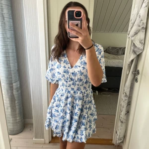 Vit och blå blommig kortklänning - Helt ny med lappen kvar, från newyorker. Superfin kortklänning med blått blommönster på vit botten. Klänningen har v-ringning, korta ärmar och smock i midjan som ger en snygg passform. Kjolen är lätt utställd och tyget känns luftigt, perfekt för varma dagar.