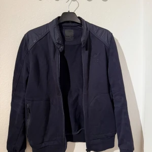 Hackett X Aston Martin jacka/tröja - Snygg marinblå bomberjacka från Hackett x Aston Martin Racing. Jackan har quiltade axlar, dragkedja framtill och diskreta loggor på bröstet och nacken. Tillverkad i bomull och polyester, perfekt för dig som gillar sportig och clean stil.