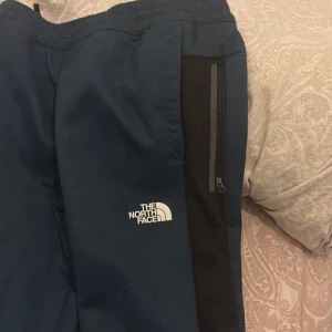 Blå träningsbyxor från The North Face - Mörkblå träningsbyxor från The North Face med svart panel längs sidorna och vit logga på låret. Byxorna har elastisk midja och dragkedjeficka på sidan. Perfekta för chill eller träning, snygg och sportig look.