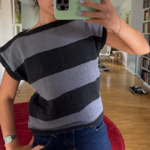 Randig stickad topp i svart och grått - Säljer en stickad topp med breda svarta och grå ränder. Modellen är ärmlös med lös passform och har ribbade kanter vid hals, ärmhål och nederkant