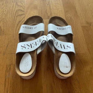 Vita sandaler från Sweeks of Sweden - Snygga vita sandaler från Sweeks of Sweden med två breda remmar i syntetmaterial och coolt utskuret logotypmönster. Fotbädden är i mjuk mocka och sulan är platt för skön komfort. Perfekta för varma dagar och chill stil.