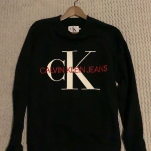 Svart sweatshirt från Calvin Klein Jeans - Svart sweatshirt från Calvin Klein Jeans med stort vitt CK-tryck och röd broderad logga på bröstet. Tröjan har rund hals, långa ärmar och är tillverkad i mjuk bomull. Perfekt för en chill och stilren look. Hör hjärna av er😁
