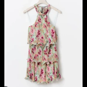 Blommig kortklänning med volanger - Supersöt kortklänning med blommigt mönster. Klänningen har hög hals, ärmlös och flera volangdetaljer. 